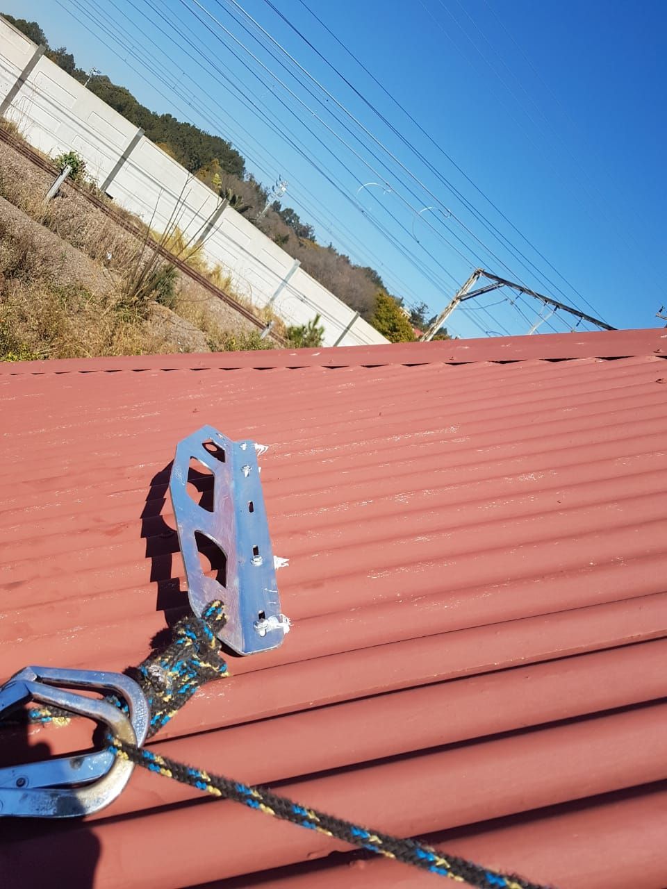 A fall arrest device secured to a red corrugated metal roof, with power lines and a hillside in the background.