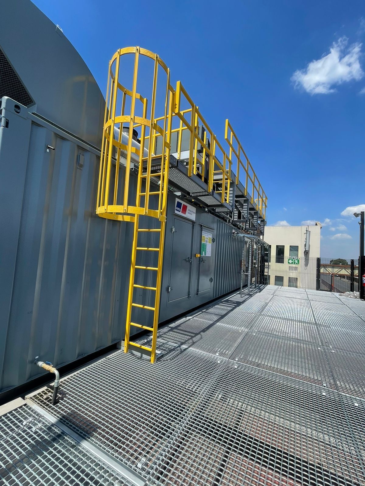 Yellow ladder and railing on a large gray rooftop unit with a patterned metal surface, blue sky in background.