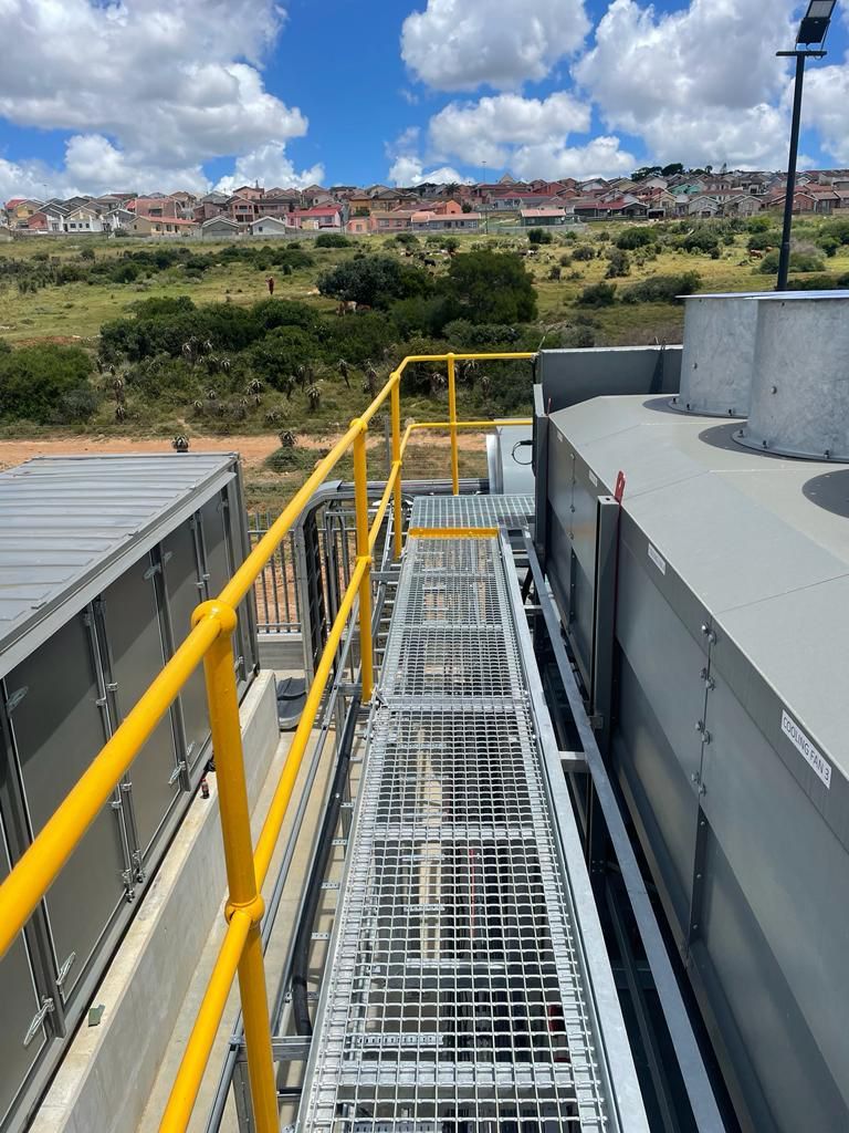 Metal walkway with yellow handrails on rooftop, overlooking landscape with houses.