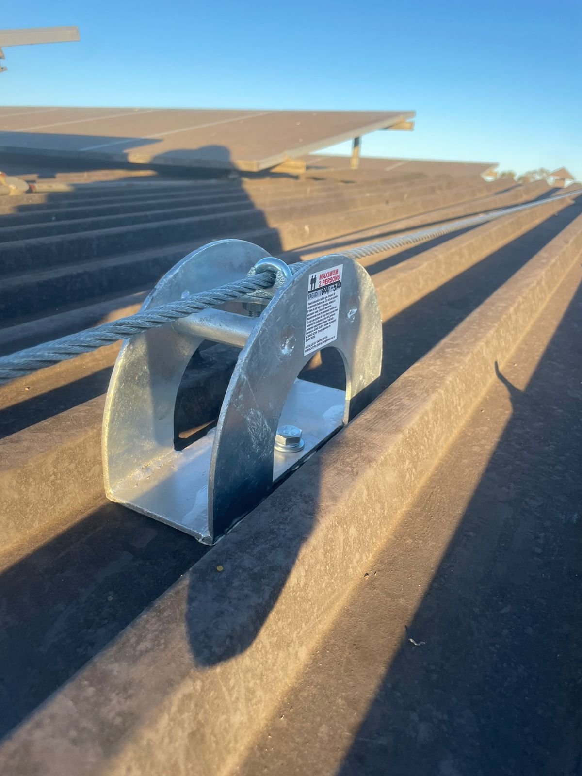 Metal cable anchor on corrugated roof with solar panels in background.