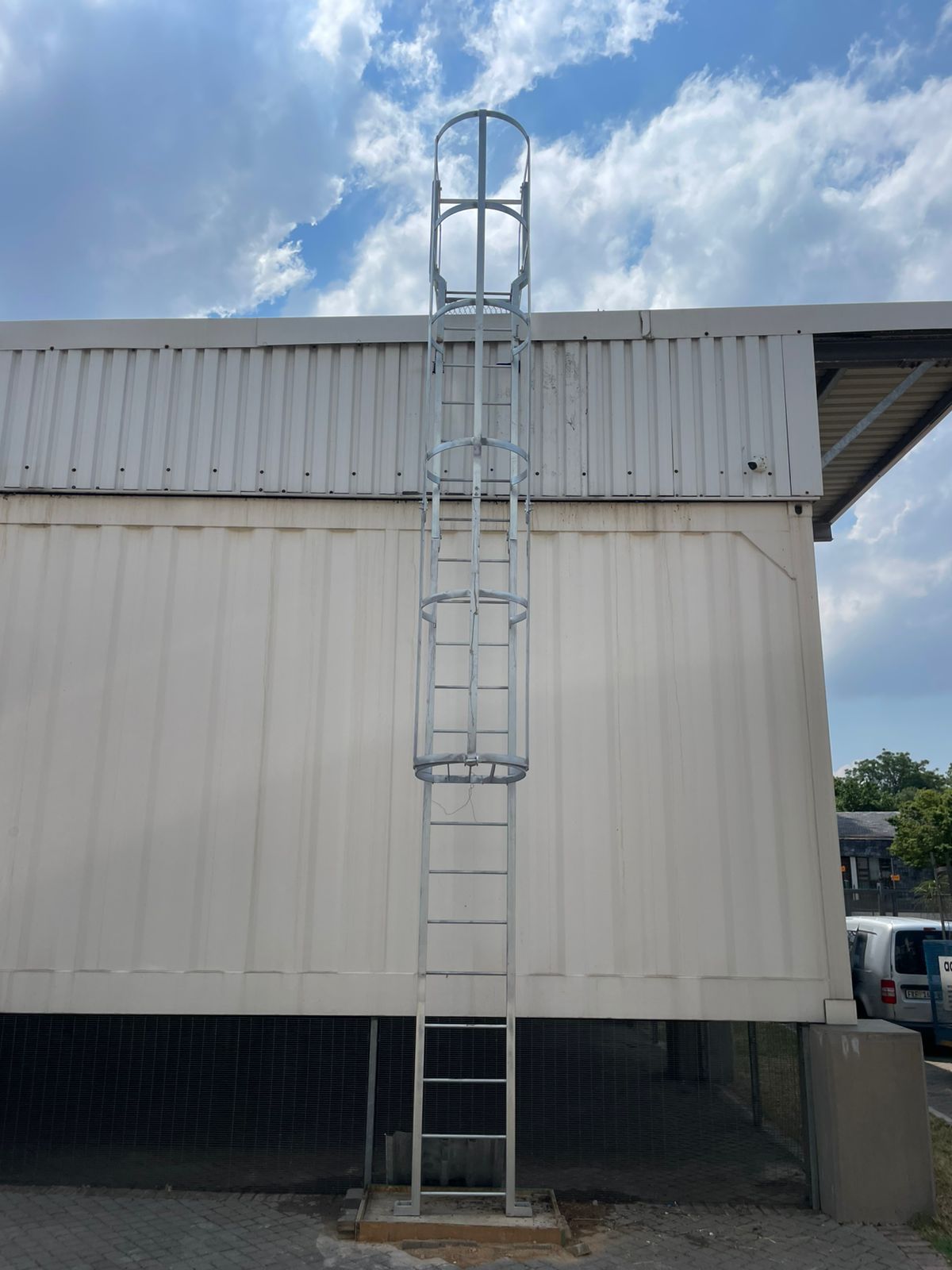 Metal ladder with safety cage attached to the side of a white building under a cloudy sky.