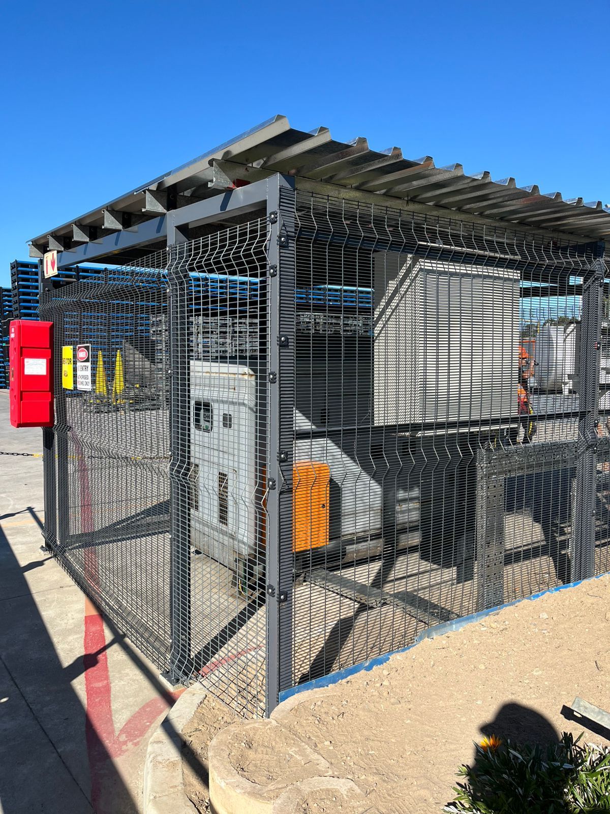 Metal electrical equipment enclosure with black fencing, overhead trellis, and red emergency box.