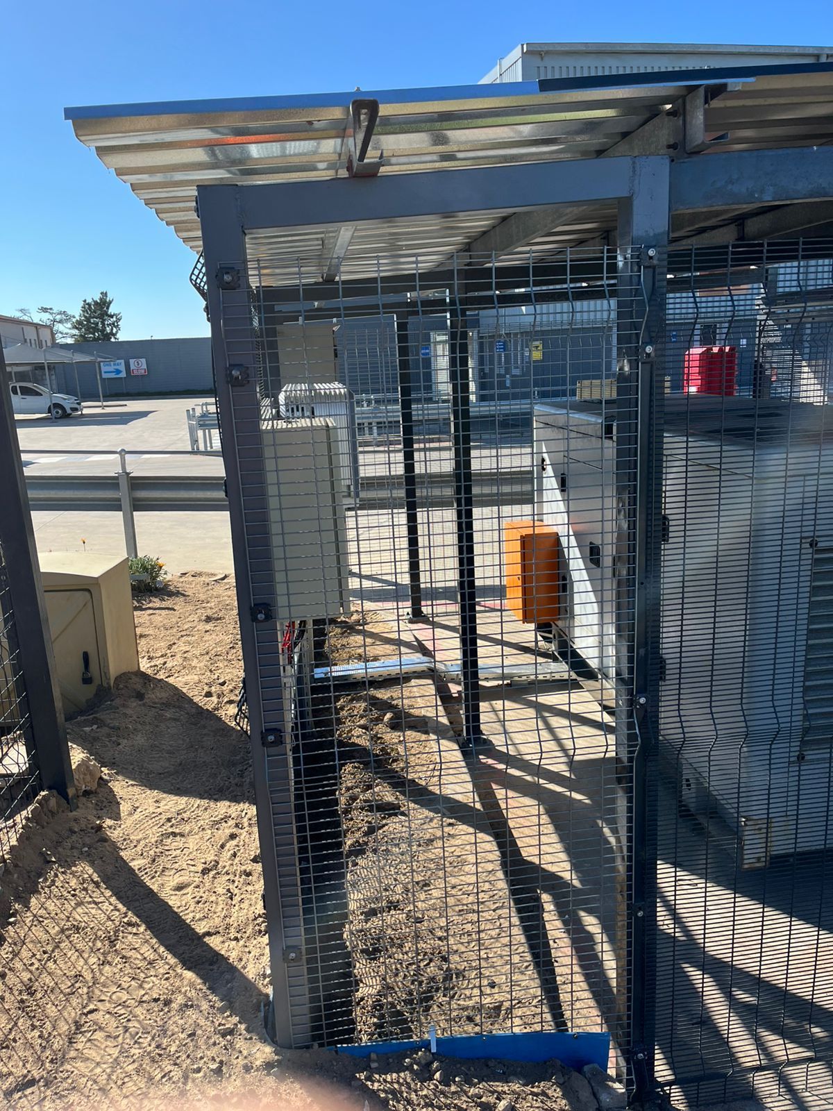 Metal fenced enclosure with electrical equipment, topped with a solar panel array, in an outdoor setting.