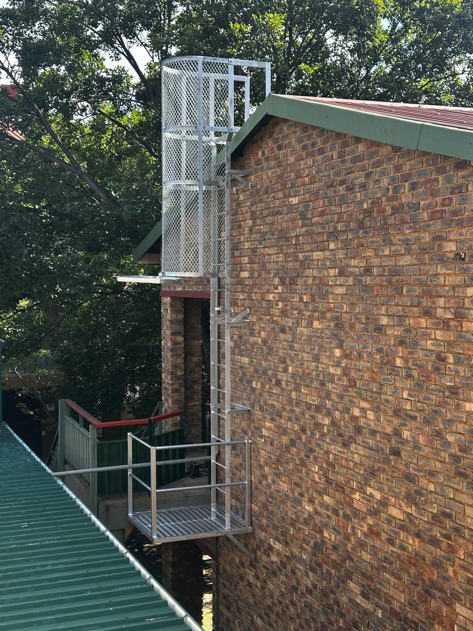 Metal fire escape attached to a brick building. Green roof visible in the foreground. Trees in the background.