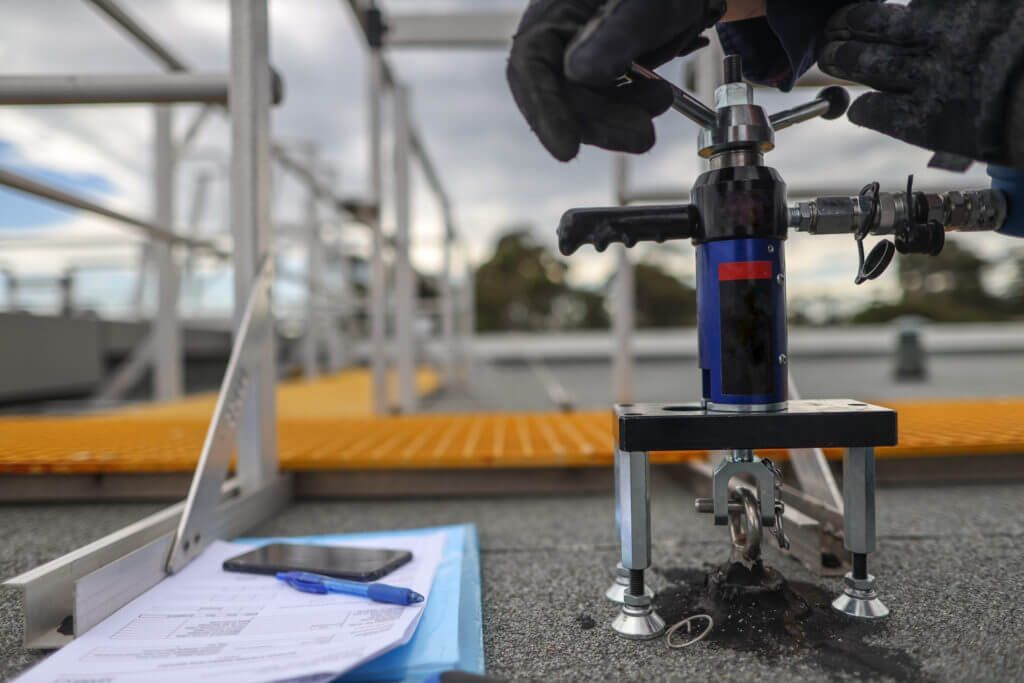 Gloved hands use a tool on a roof. A clipboard and scaffolding are in the blurry background.