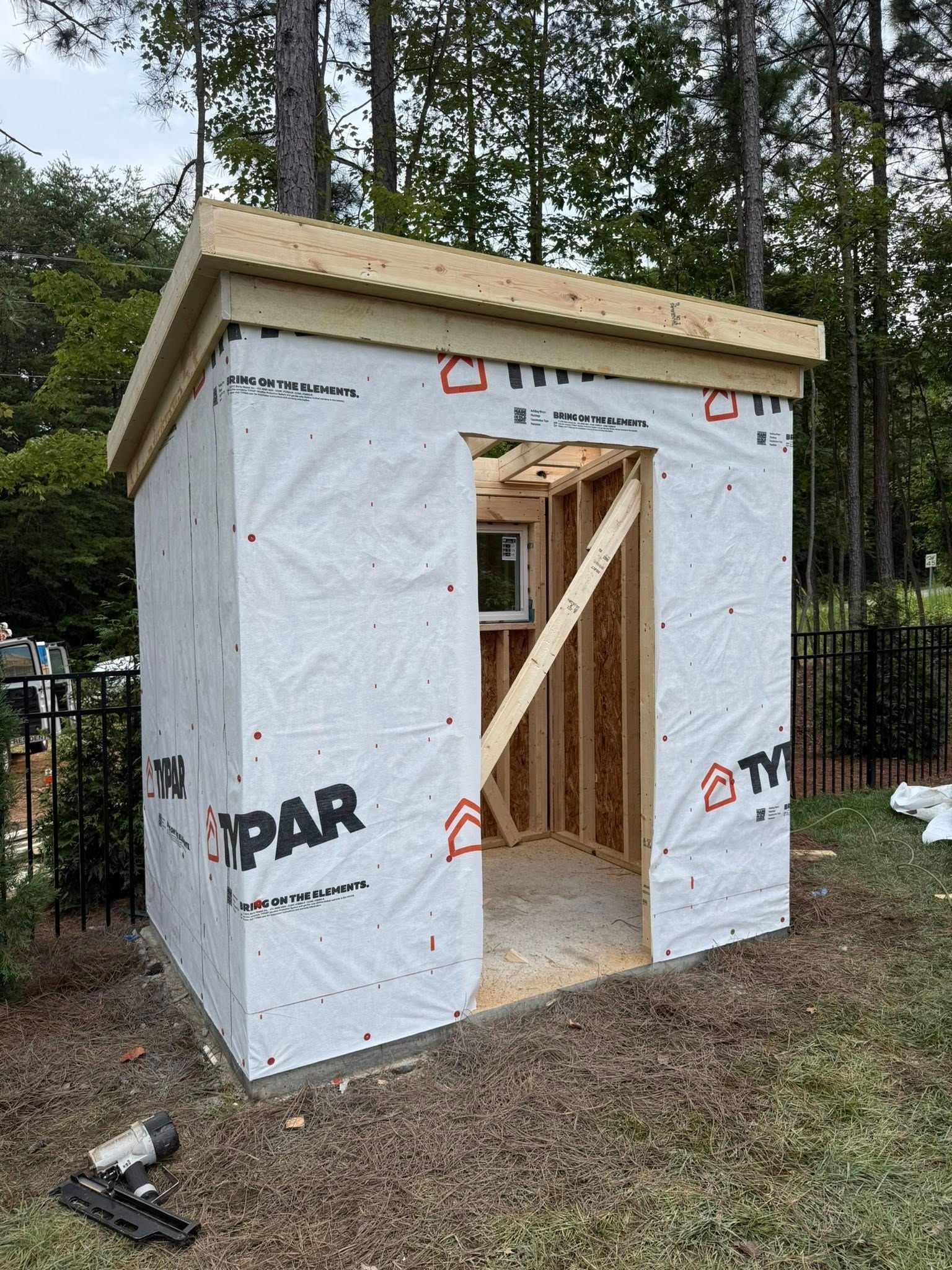 Small, unfinished shed with open doorway and window, covered in white Typar and topped with wood roof.