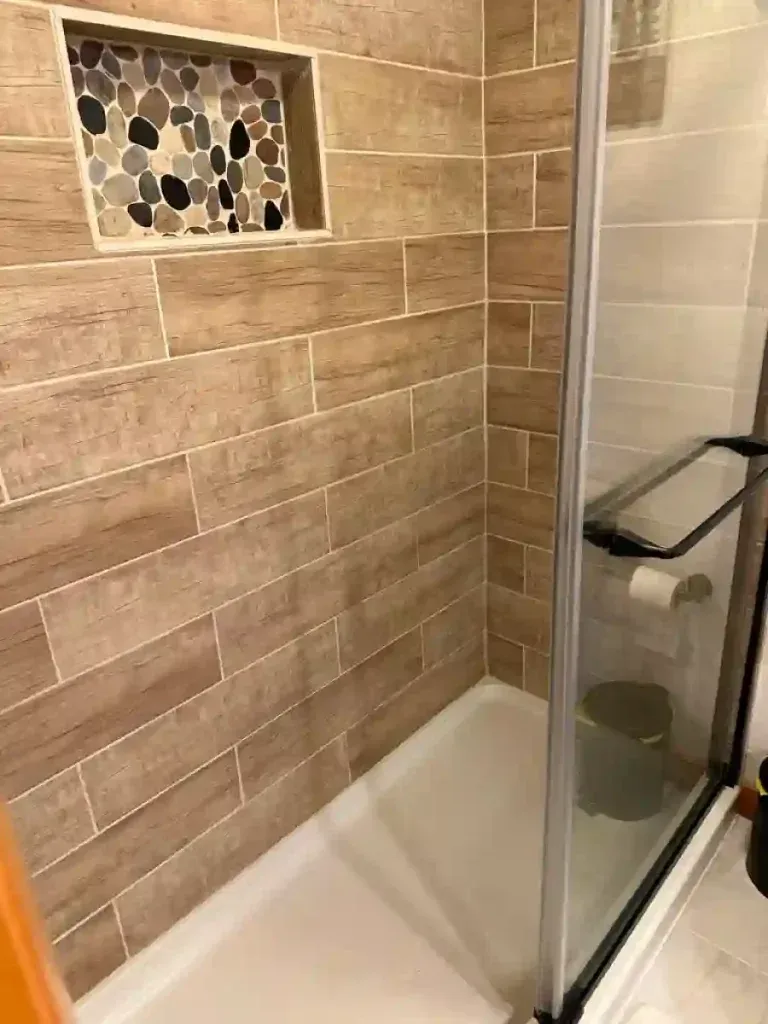 Shower stall with light brown wood-look tiles, a pebble-tiled alcove, and a glass door.