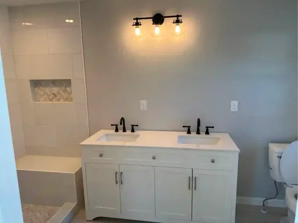 Bathroom with white vanity, double sinks, and black fixtures. Gray walls, shower with built-in bench.