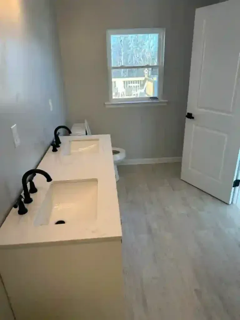 Bathroom with white vanity, black faucets, toilet, and a window. Light wood-look flooring. White door.