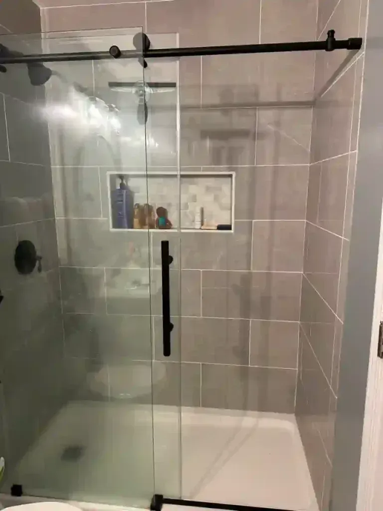 Shower with clear glass door, black hardware, gray tiled walls, recessed shelf with toiletries.