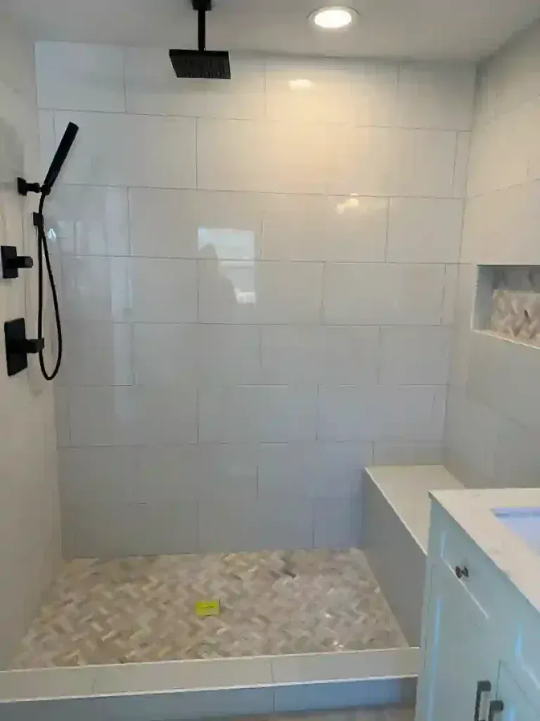 White-tiled shower with black fixtures, built-in seat, and herringbone floor. Recessed shelf on the right.