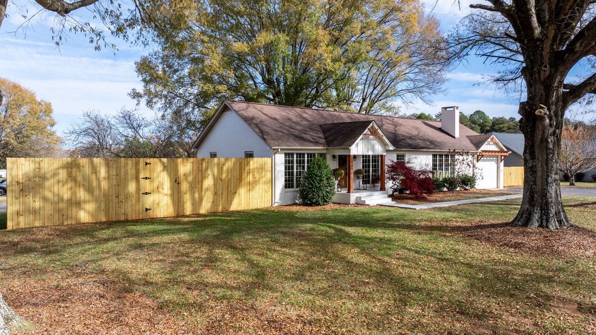 Cottage-style home with white siding, brown roof, and wooden fence in front yard with autumn leaves.