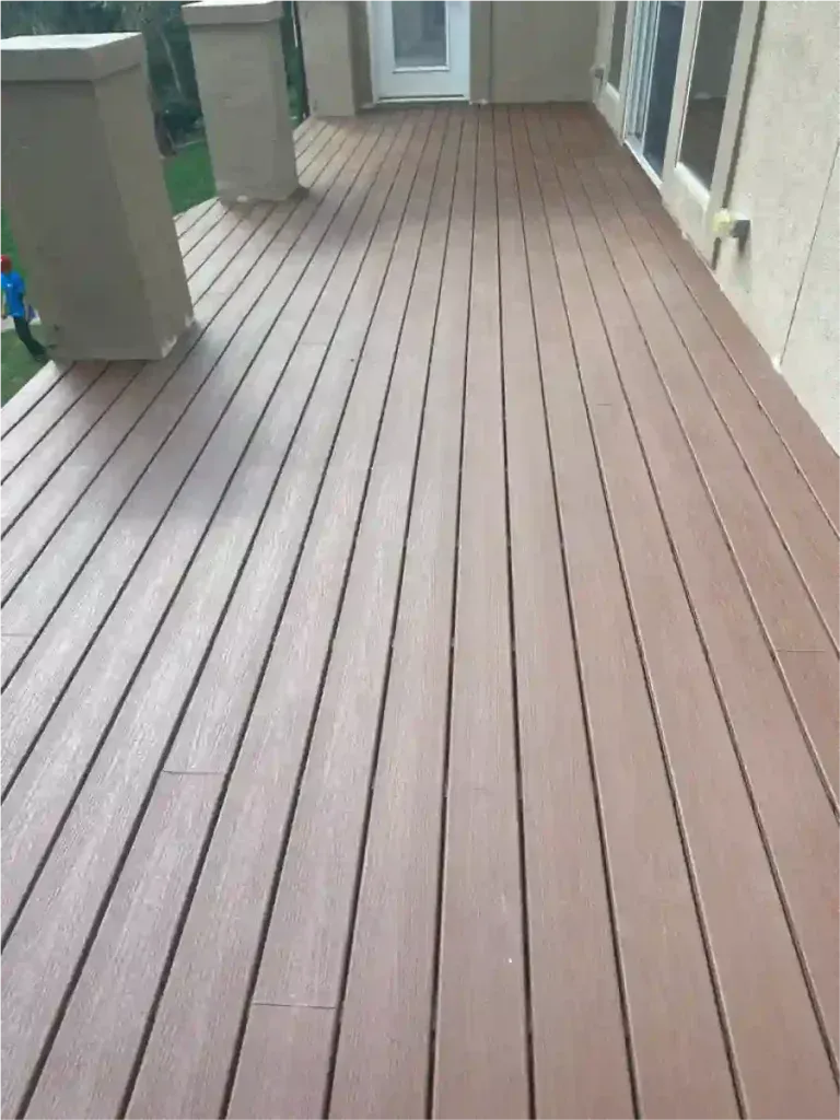 Wooden deck with reddish-brown planks extends from a house, with a white door and green lawn visible.