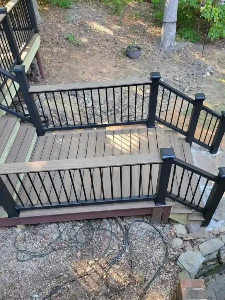 Deck with brown composite decking, black railings, and steps leading down to a wooded area.