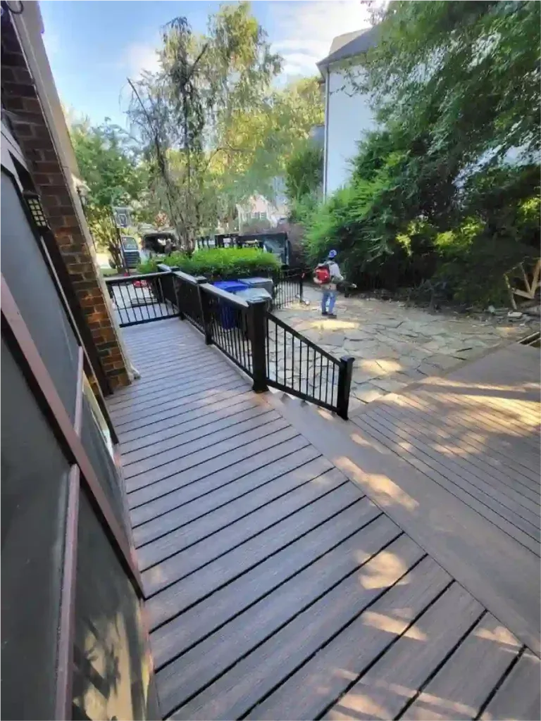 Wooden deck with black railings. A paved area extends beyond, surrounded by trees and a building.