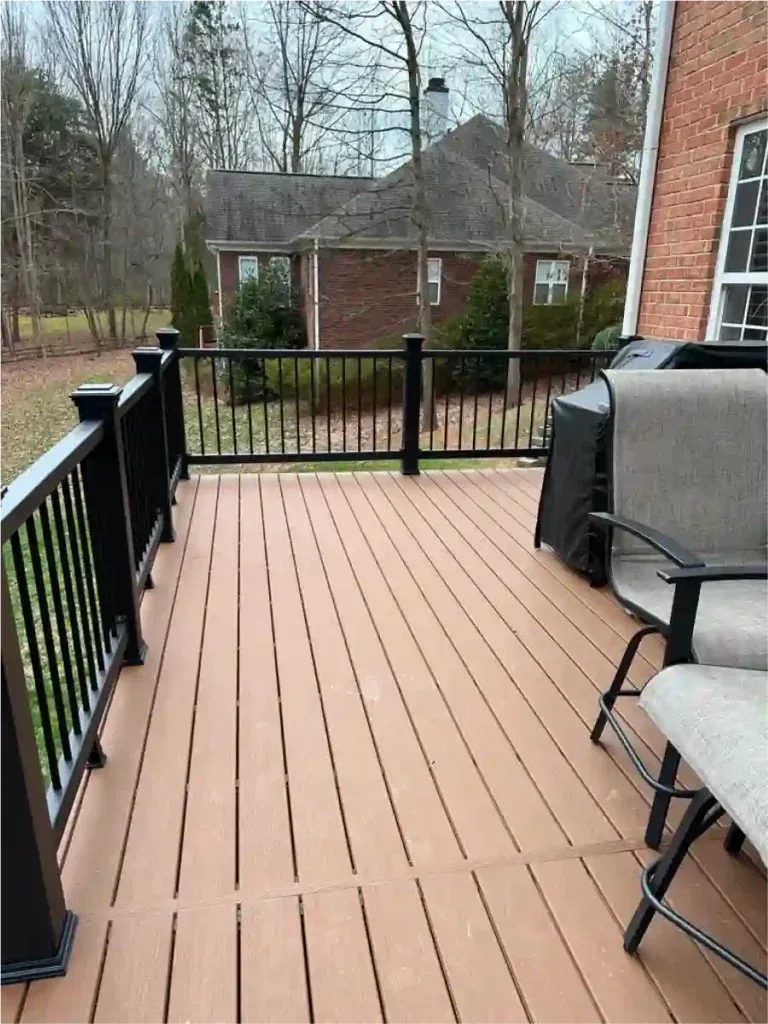 Wooden deck with black railing, overlooking a yard and a brick house with a brown roof.
