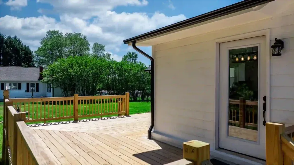 Wooden deck with railing, white house, glass door, and green lawn.