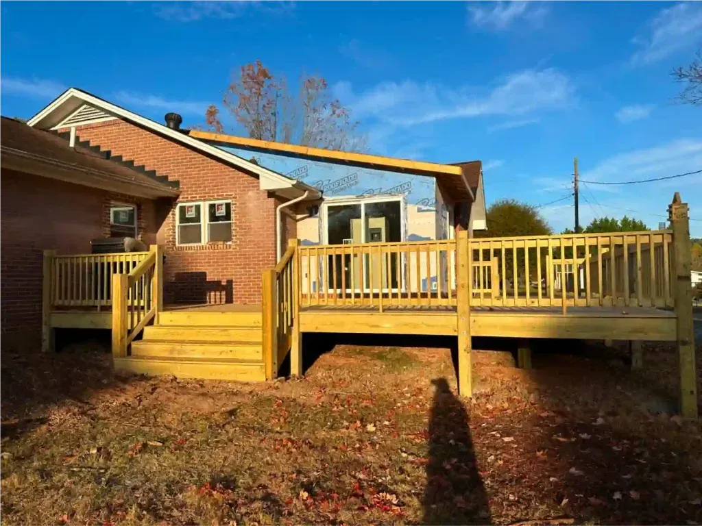 Wooden deck attached to a brick house, featuring steps, railing, and a small pergola under a clear blue sky.