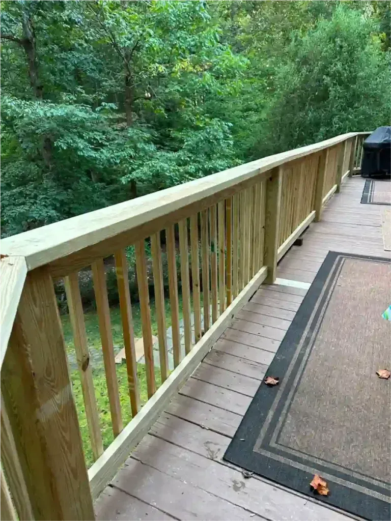 Wooden deck railing overlooking a wooded area. The deck has a rug, with a grill visible in the background.
