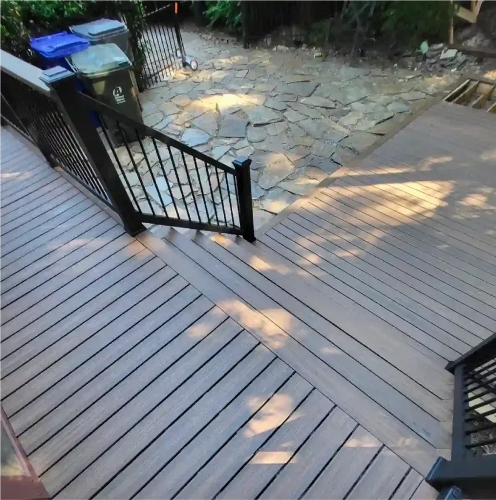 Wooden deck with black railing and steps leading to a flagstone pathway, surrounded by greenery.