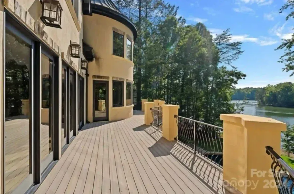 Exterior deck overlooking a lake, beige stucco house with black framed windows, ornate black railing, and lush trees.