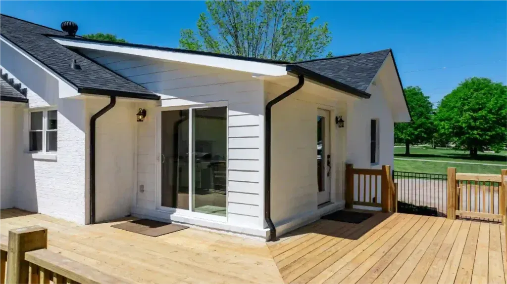 White house with black roof, wooden deck, glass sliding doors, sunny day, green field.