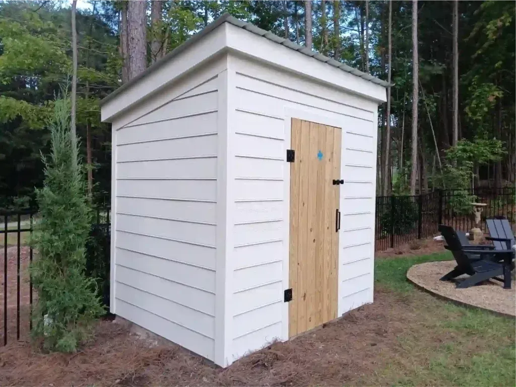 Man framing a small shed, standing on a ladder. The shed has a partially constructed wooden frame and plywood sheathing.