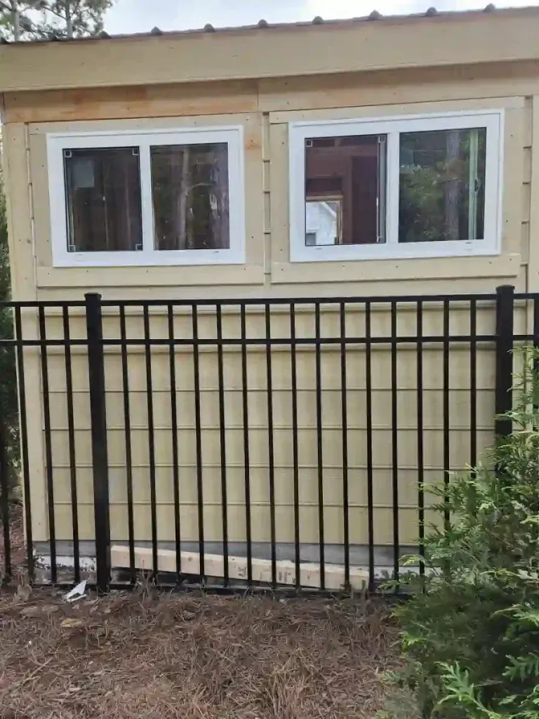 Tan building with two windows above a black metal fence, in a wooded setting.