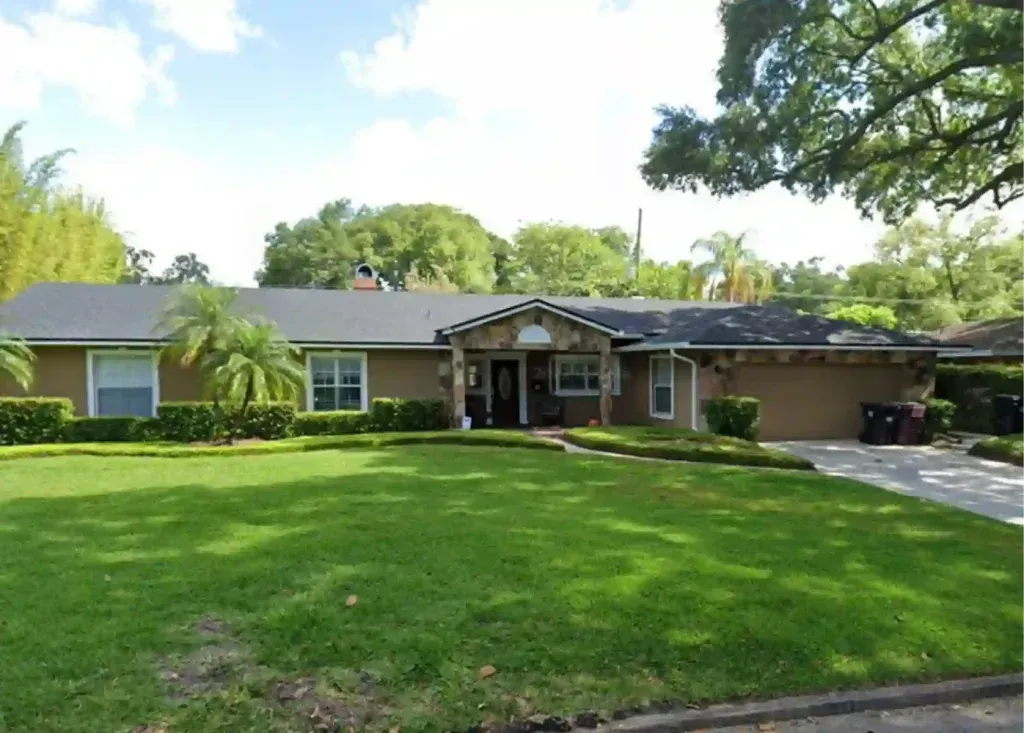 Tan, single-story house with a stone front entrance, green lawn, and a driveway.