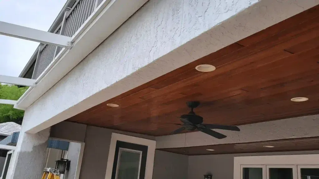 View of a covered patio ceiling with wood panels, recessed lights, and a ceiling fan; white trim.