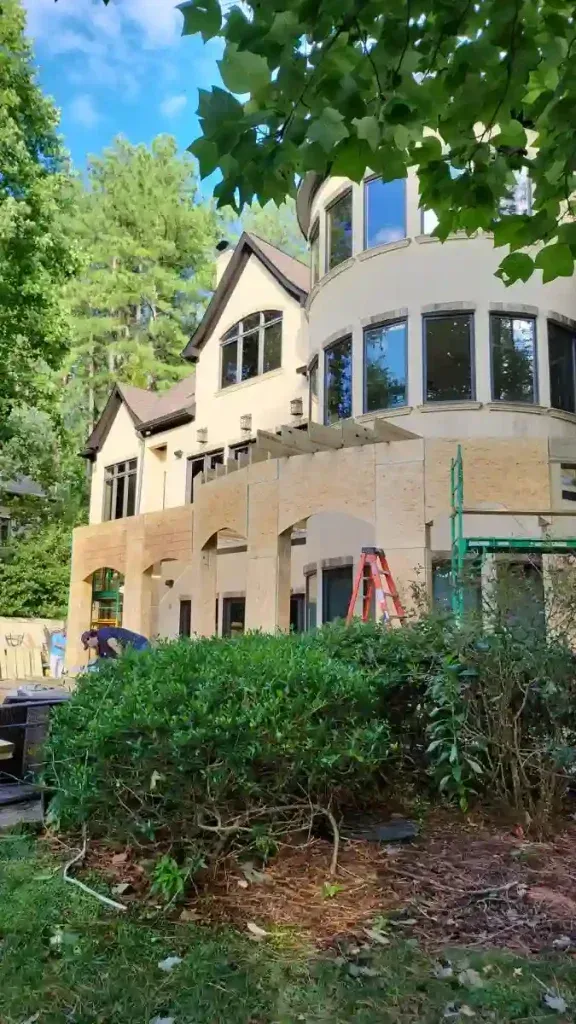 House exterior under construction, tan facade with open area; red ladder, green scaffolding, greenery.