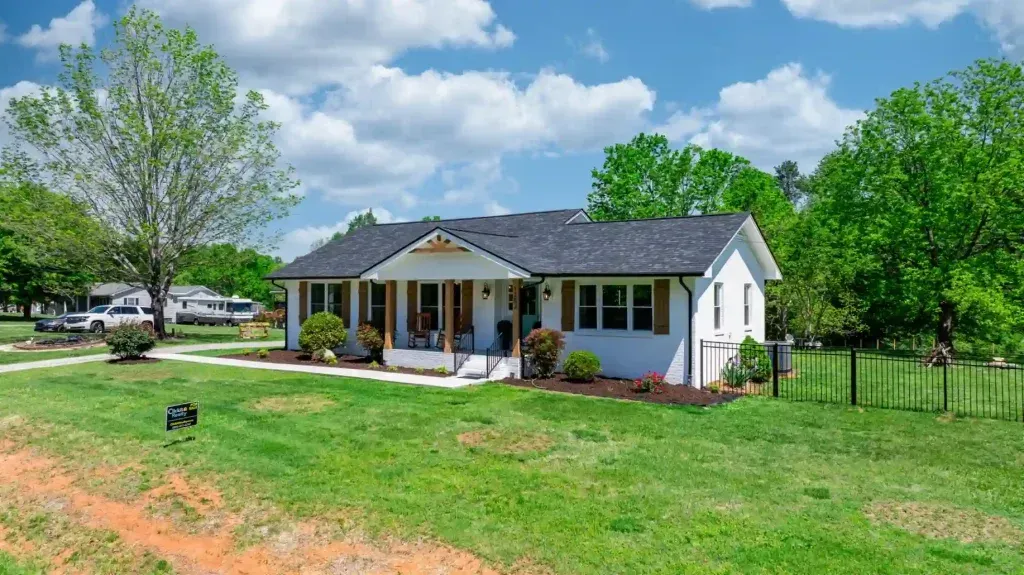 White house with porch, green lawn, trees, blue sky with clouds.