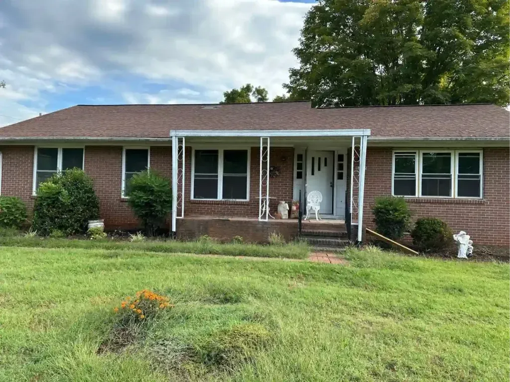 Brick ranch home with brown roof, white porch and front door, and overgrown yard.