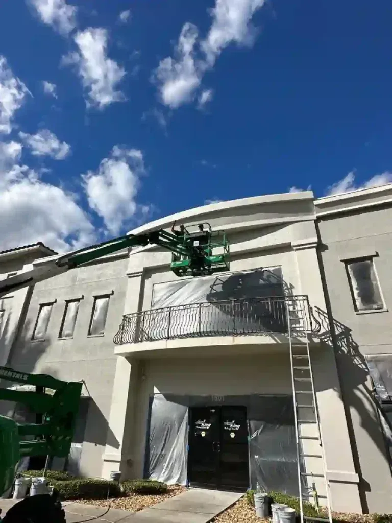 White two-story building with dark windows, palm trees, and blue sky. Sidewalk and sign in front.