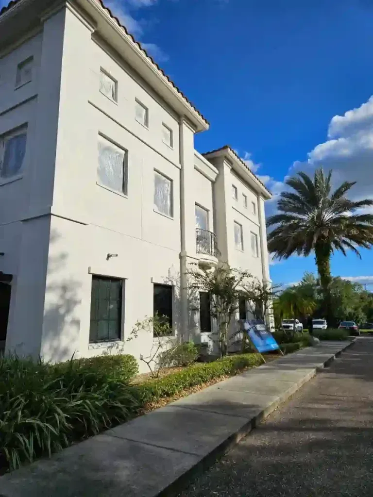 White stucco building with windows, sidewalk, palm tree, and blue sky.