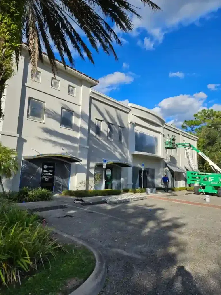 Exterior of a light grey building under renovation. A worker in a lift paints the upper facade on a sunny day.