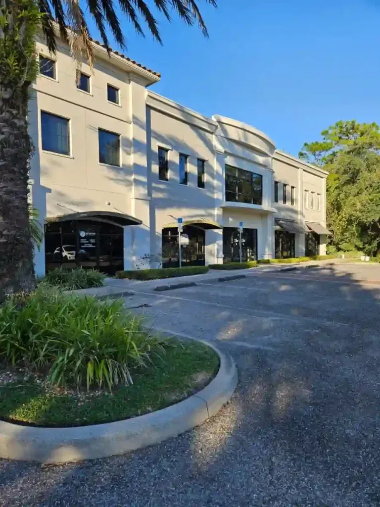 Cream-colored two-story building with dark windows and a curved roof section, surrounded by pavement and greenery.