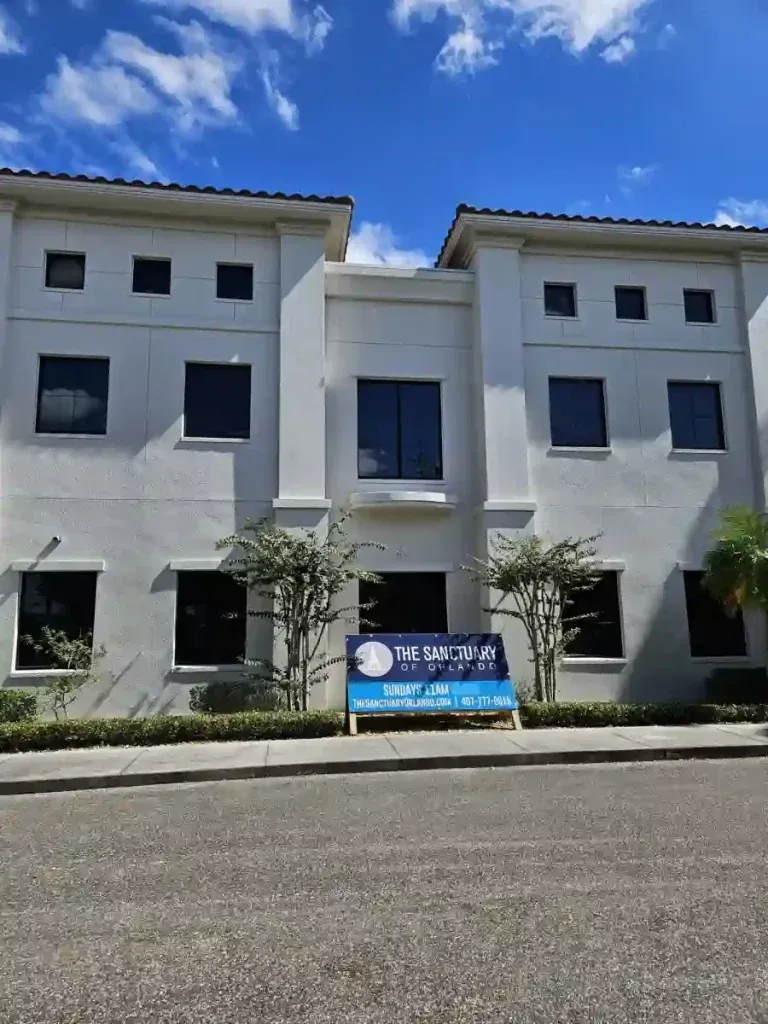 White two-story building with dark windows and a sign in front; blue sky with clouds.
