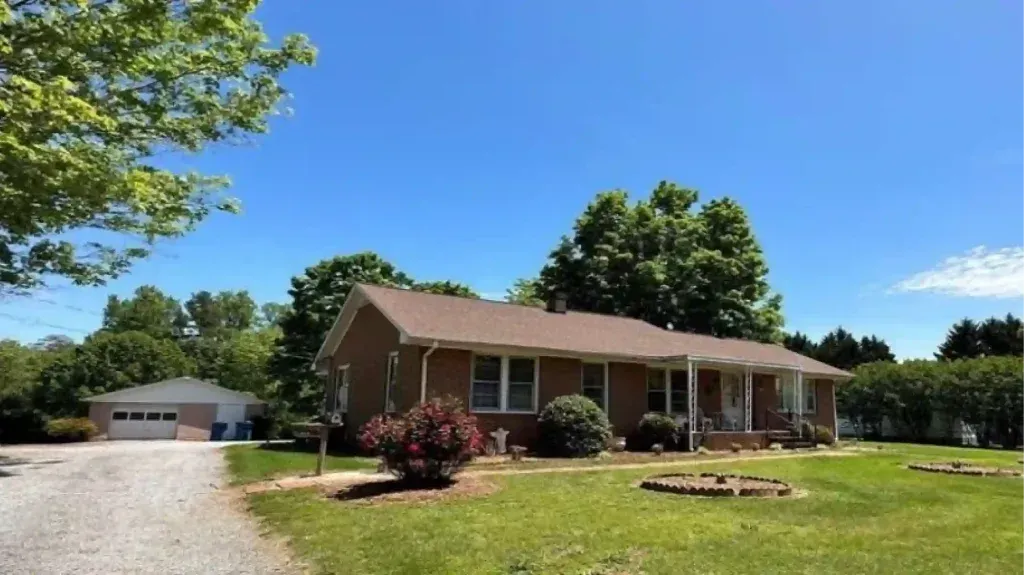A one-story brown house with a porch and a detached garage on a grassy lot under a blue sky.