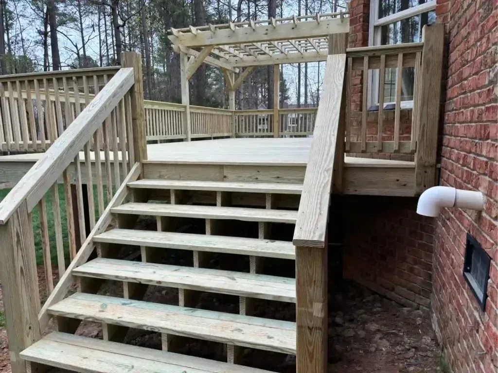 Wooden deck with stairs, railing, and pergola, attached to a brick house in a wooded area.
