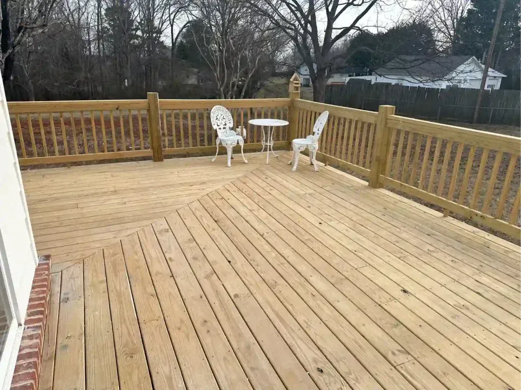 Wooden deck with railing, two white chairs, and a small table in a backyard.