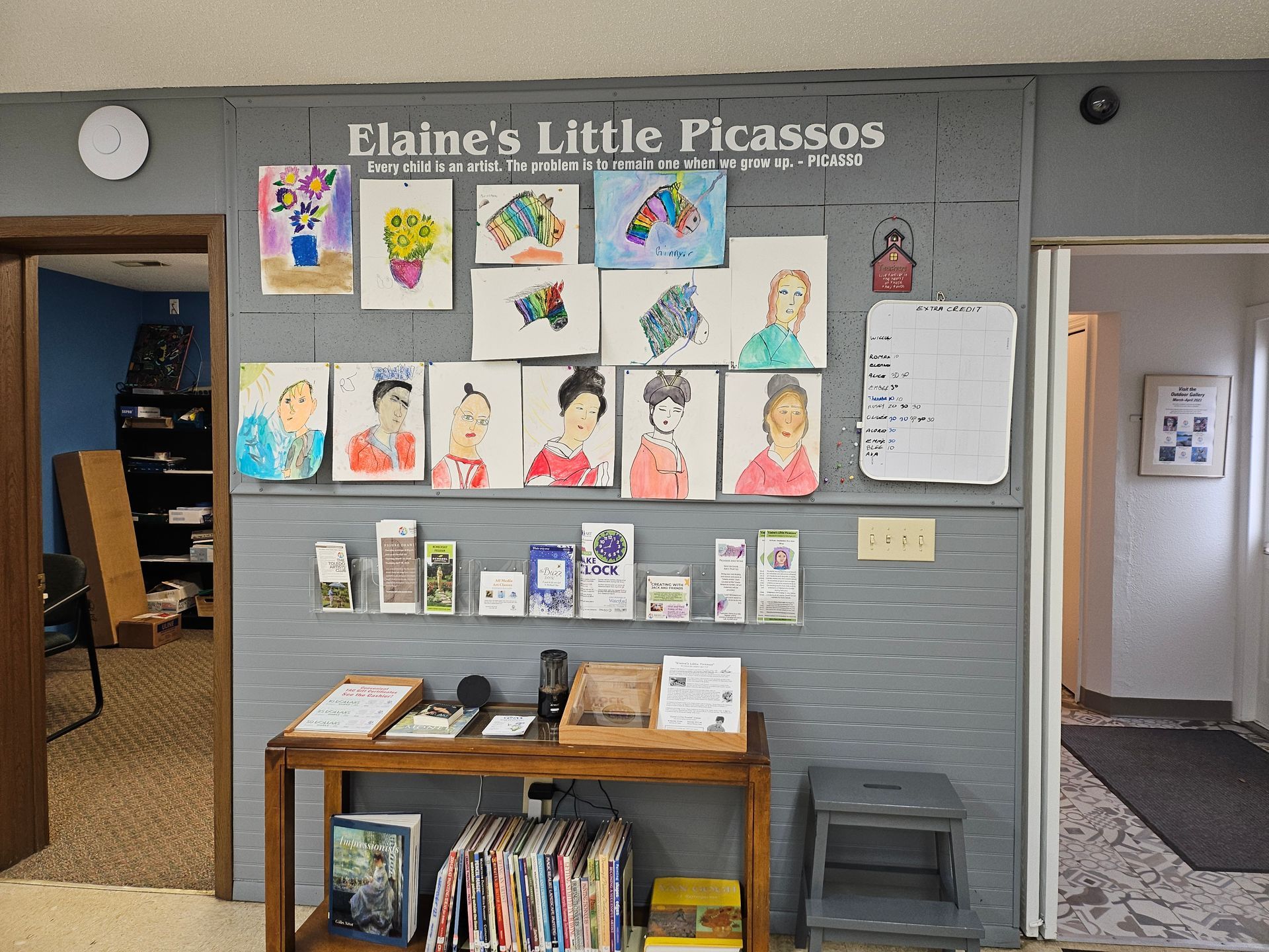 A wall with paintings on it and a sign that says elaine 's little picasso
