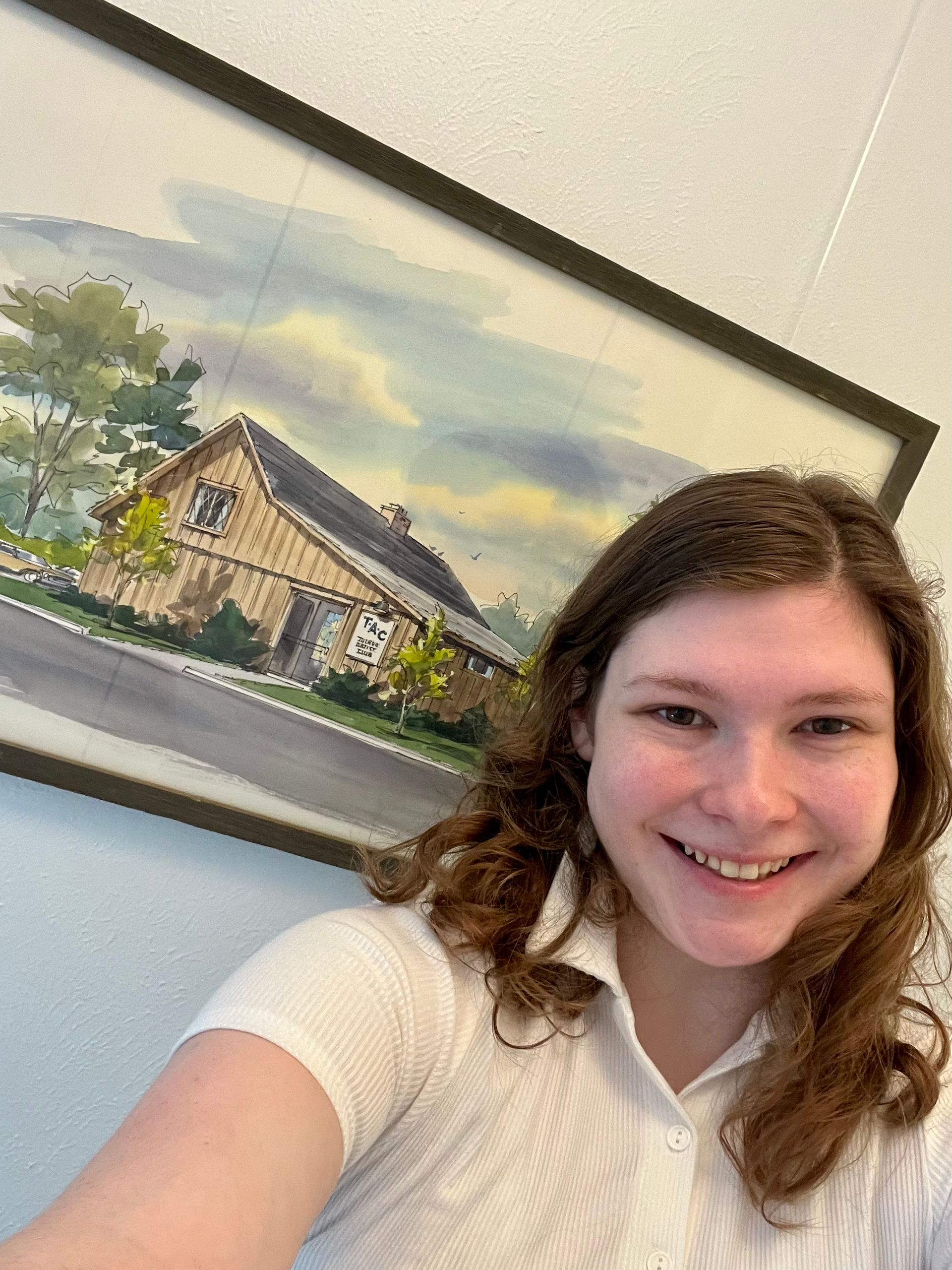 A woman is taking a selfie in front of a painting of a house.