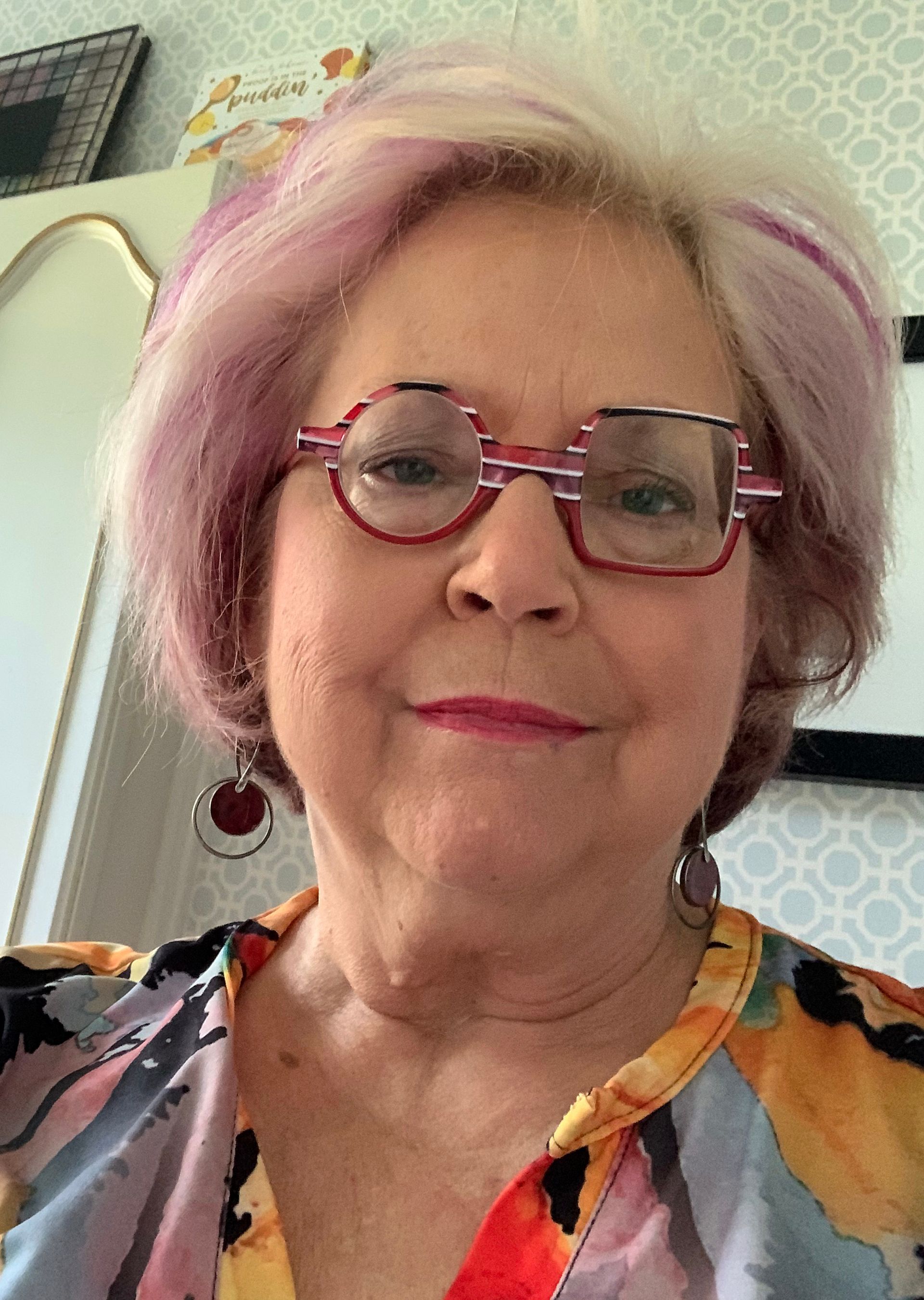 A woman with pink hair and glasses is wearing a colorful shirt.