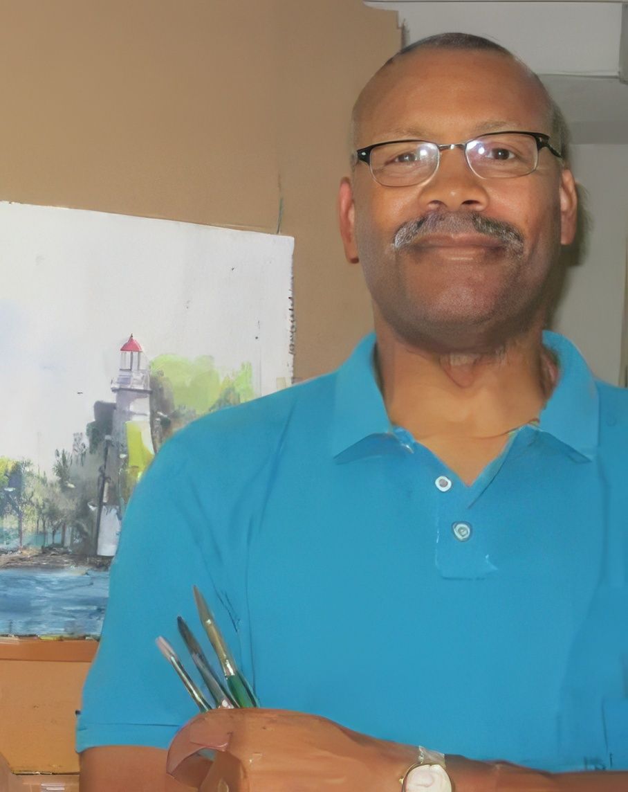 A man in a blue shirt is holding brushes in front of a painting of a lighthouse