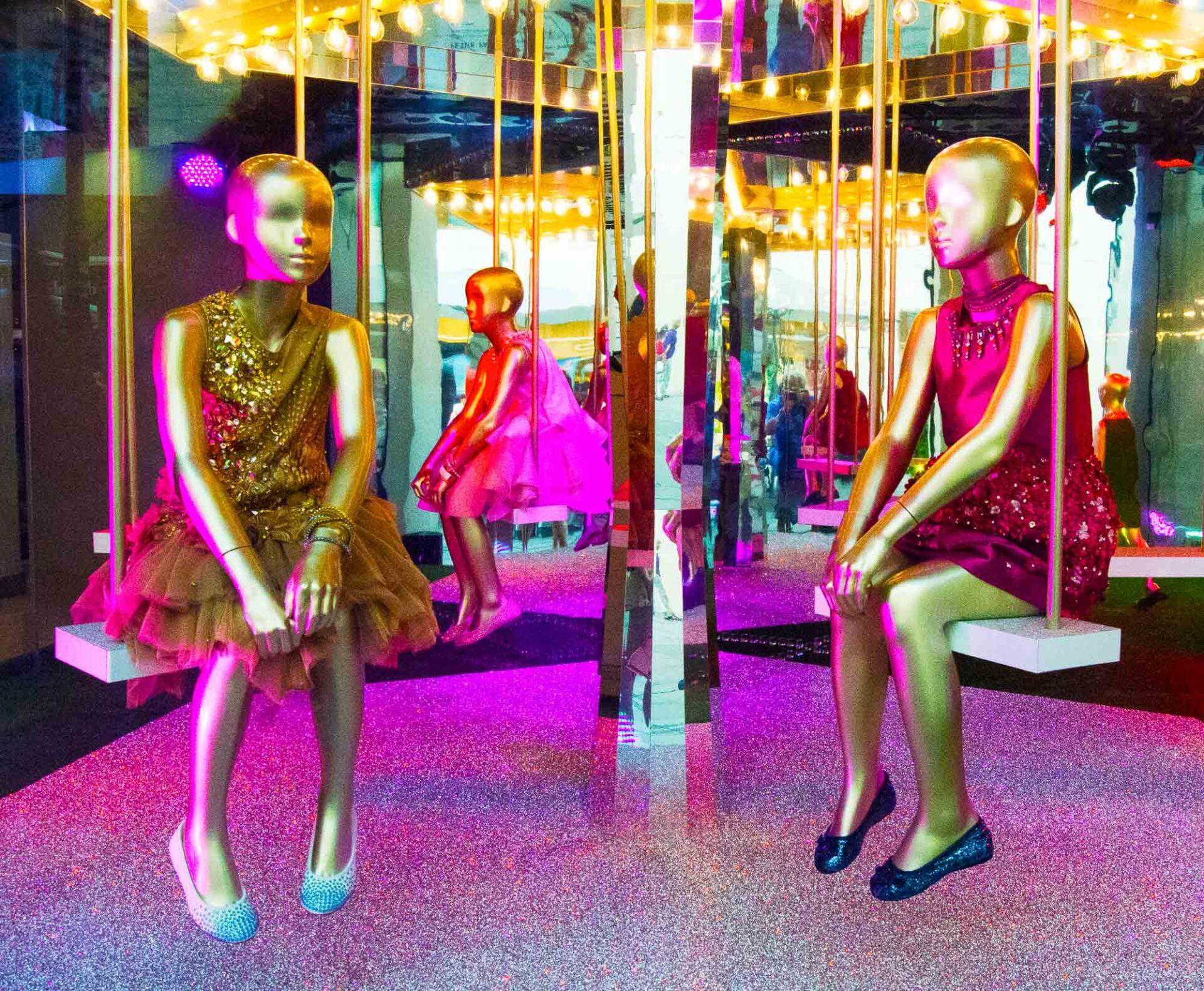 Two mannequins are sitting on swings in front of a merry go round