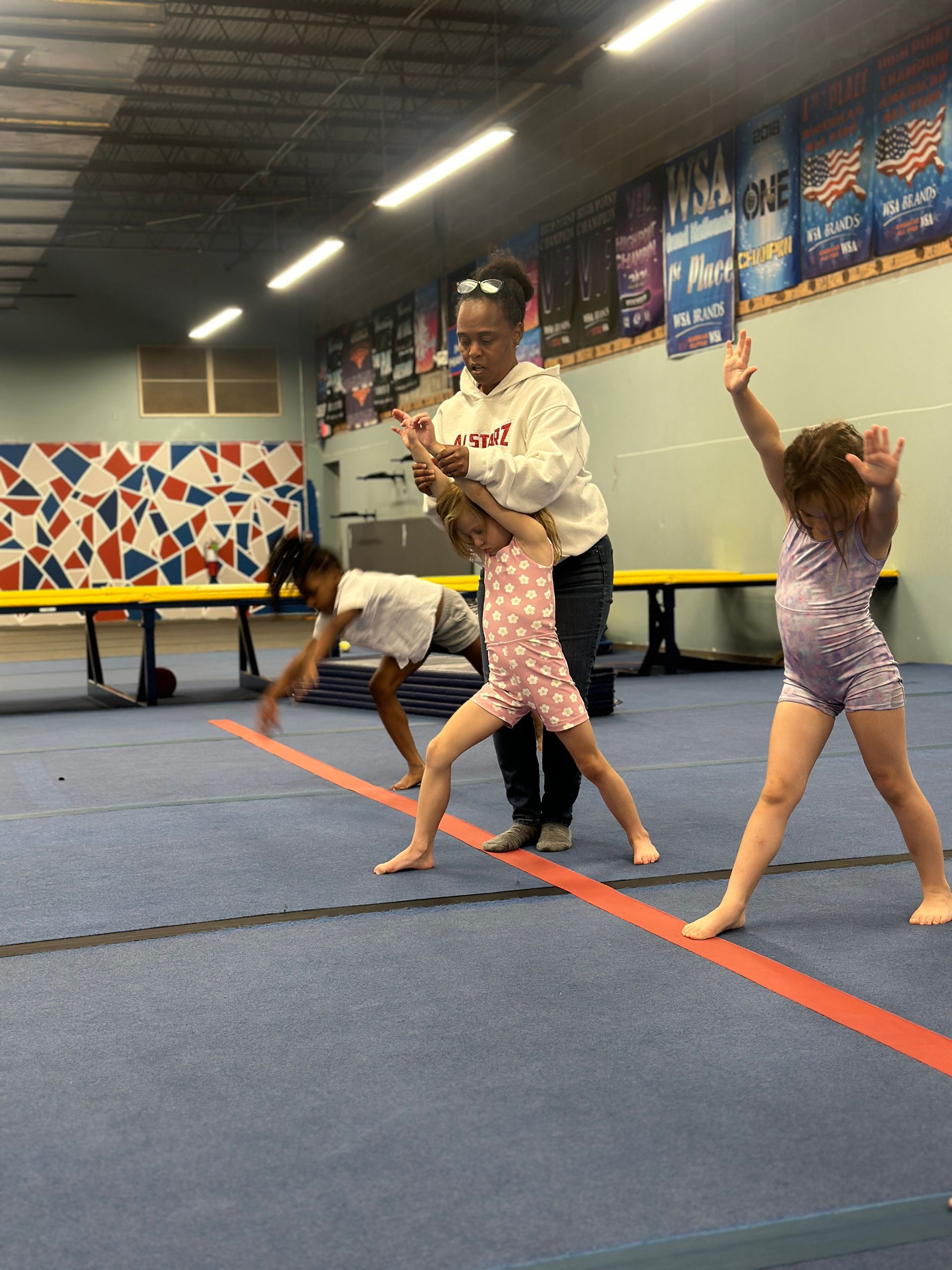 A woman is helping two young girls do exercises on a balance beam in a gym.