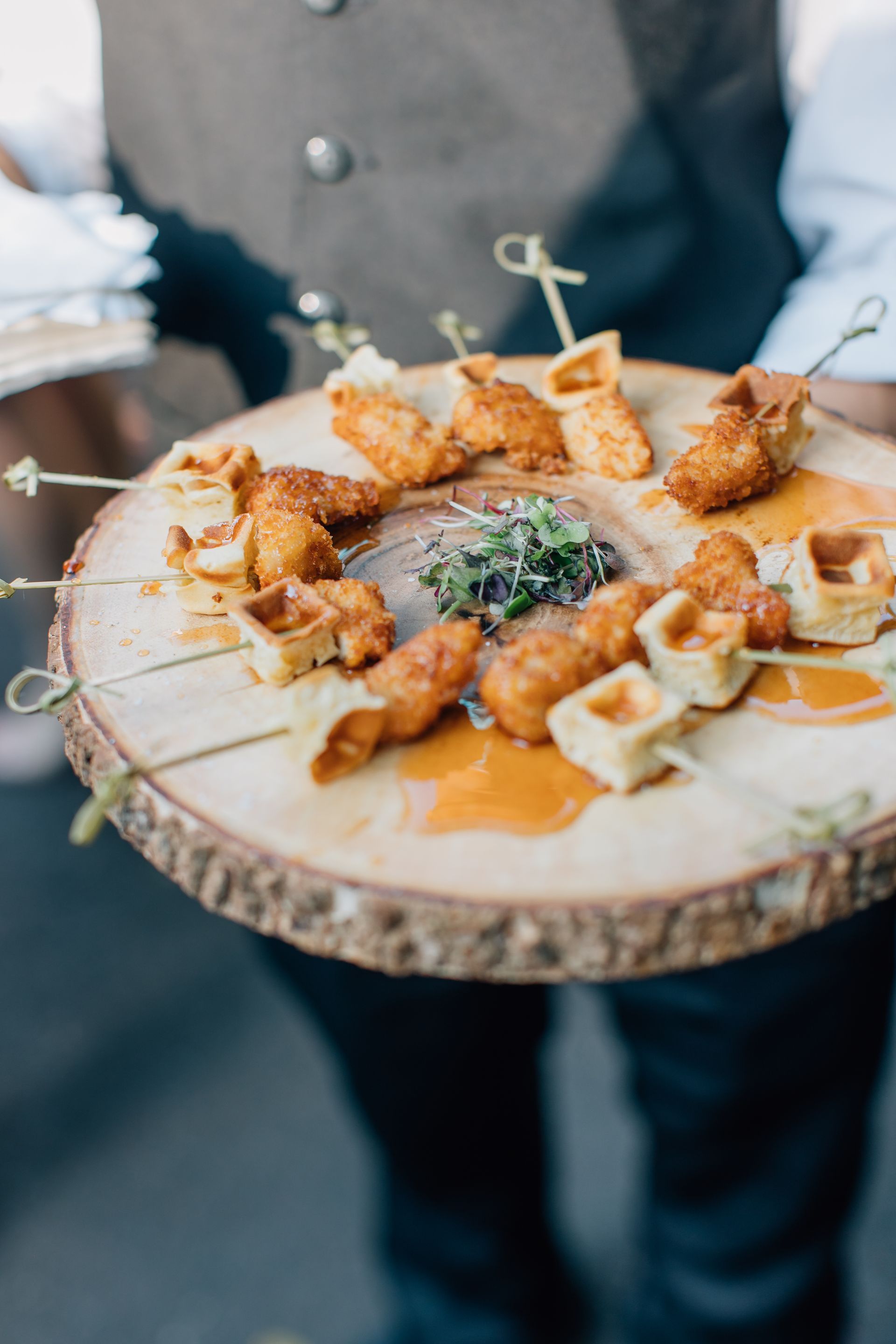 A person is holding a wooden tray of food on skewers.