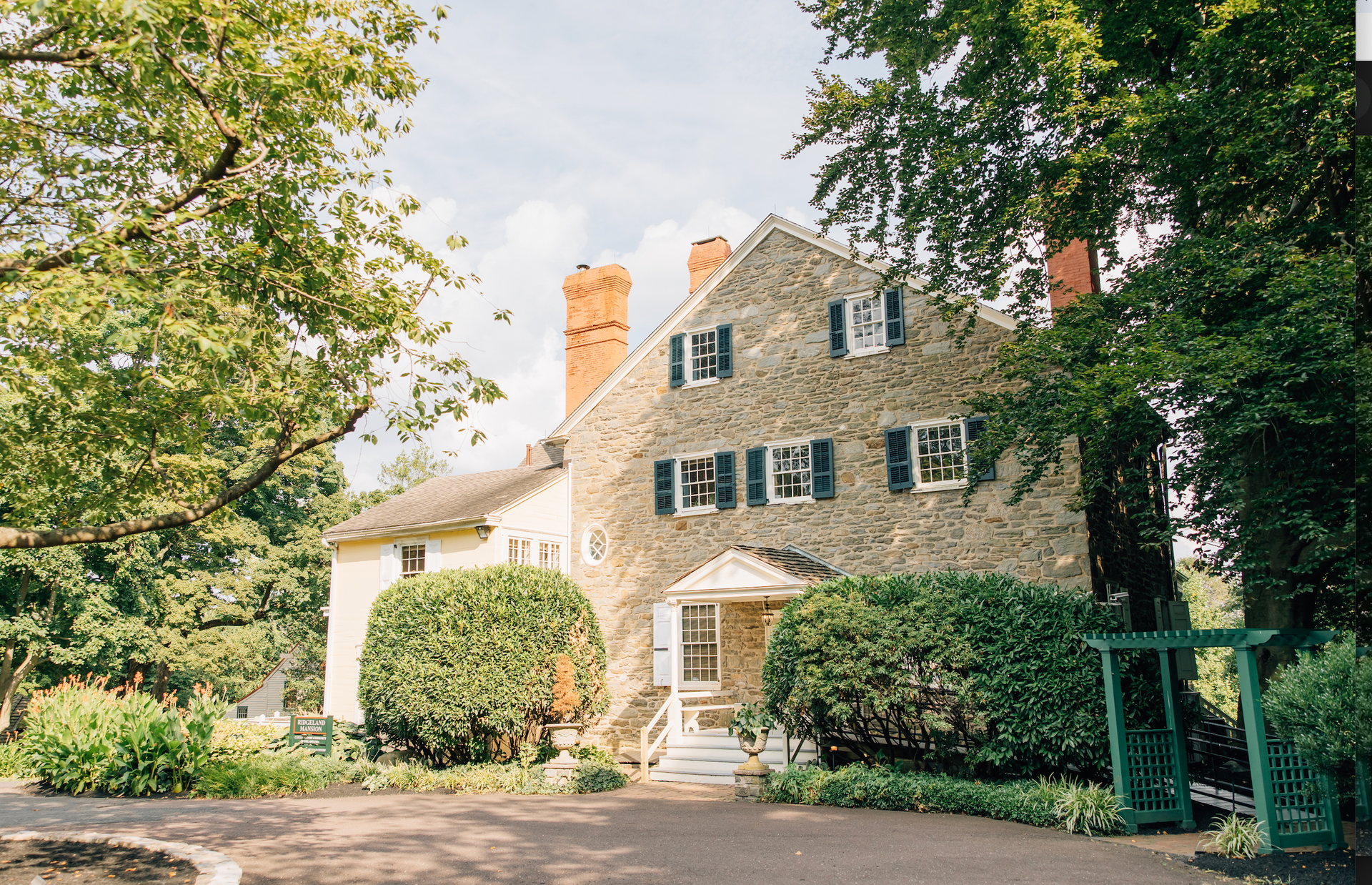 A large stone house with blue shutters is surrounded by trees and bushes.