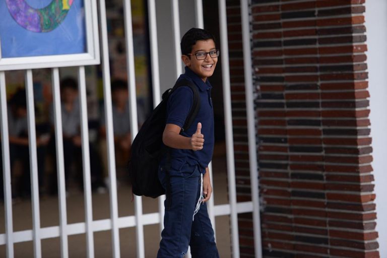 A young boy with a backpack is giving a thumbs up while walking through a school hallway.
